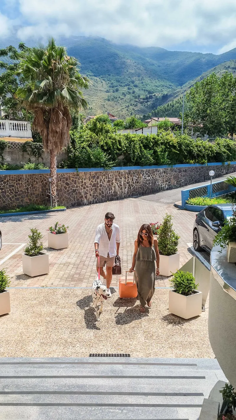Coppia con valigie e cane nel vialetto dell'hotel, con le montagne sullo sfondo